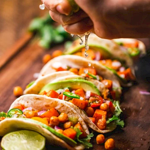 A hand squeezes lime over tacos filled with chickpeas, avocado, and vegetables, garnished with cilantro, on a wooden board.