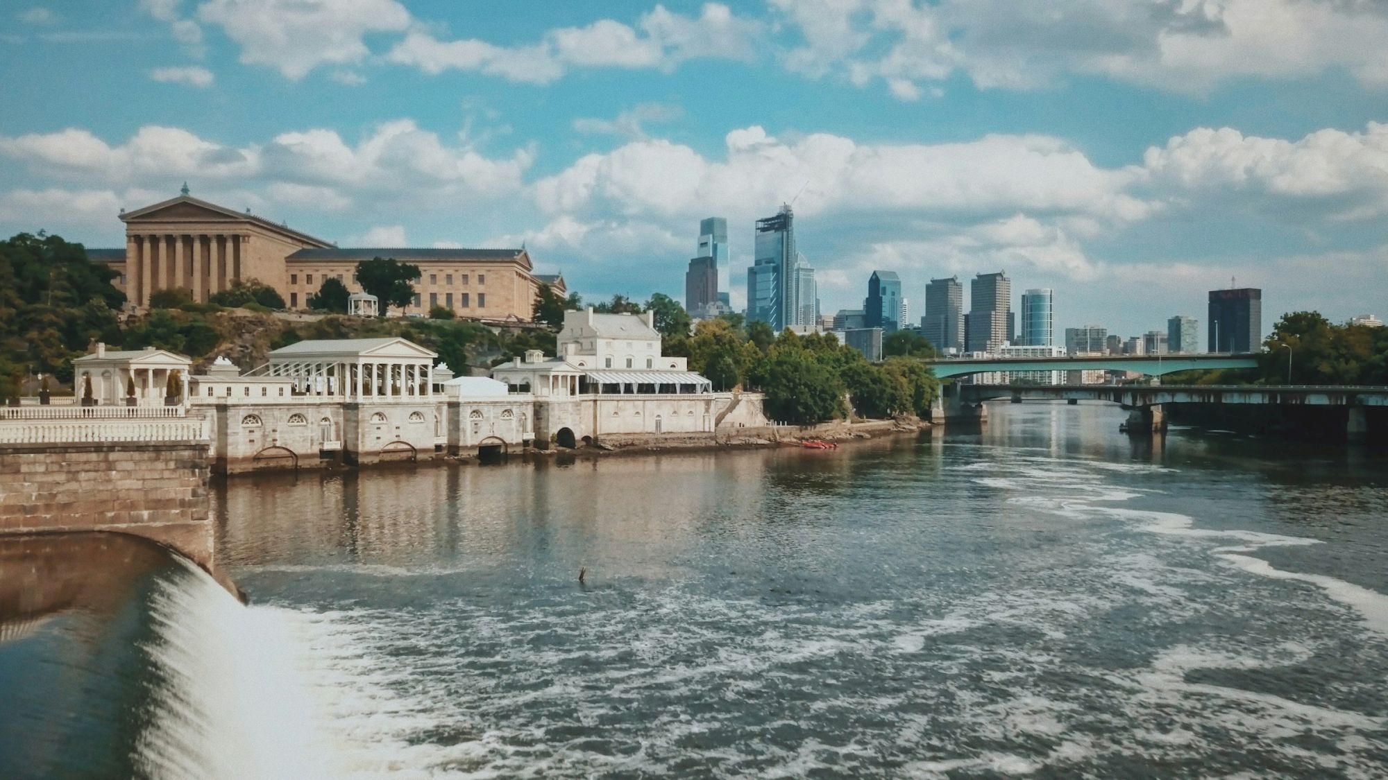 A river flows through a cityscape with classic and modern architecture, including a prominent museum building and a skyline backdrop.