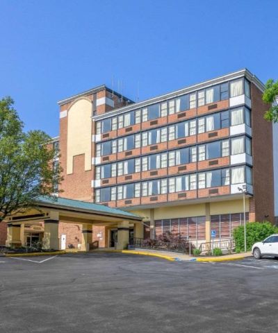The image shows a multi-story building with large windows, a parking lot, a white car, trees, and a clear blue sky in the background.