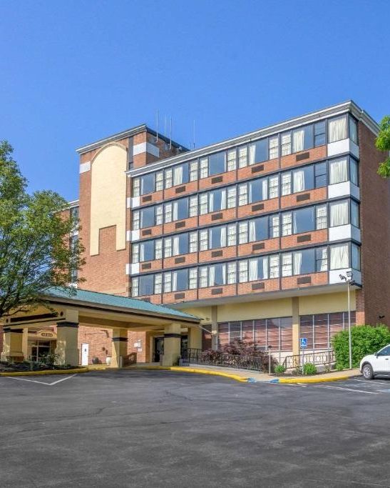The image shows a multi-story building with large windows, a parking lot, a white car, trees, and a clear blue sky in the background.