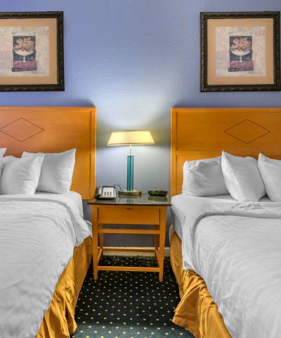 The image shows a hotel room with two double beds, a nightstand with a lamp, and two framed artworks on the wall above the beds.