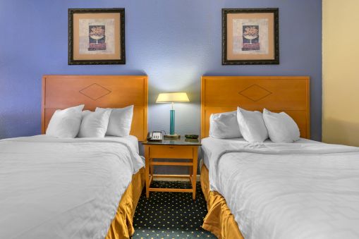 The image shows a hotel room with two double beds, a nightstand with a lamp, and two framed artworks on the wall above the beds.