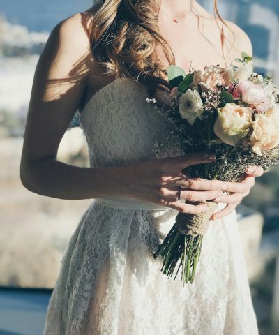 A bride in a lace wedding dress holding a bouquet of flowers, with a blurred outdoor background visible.
