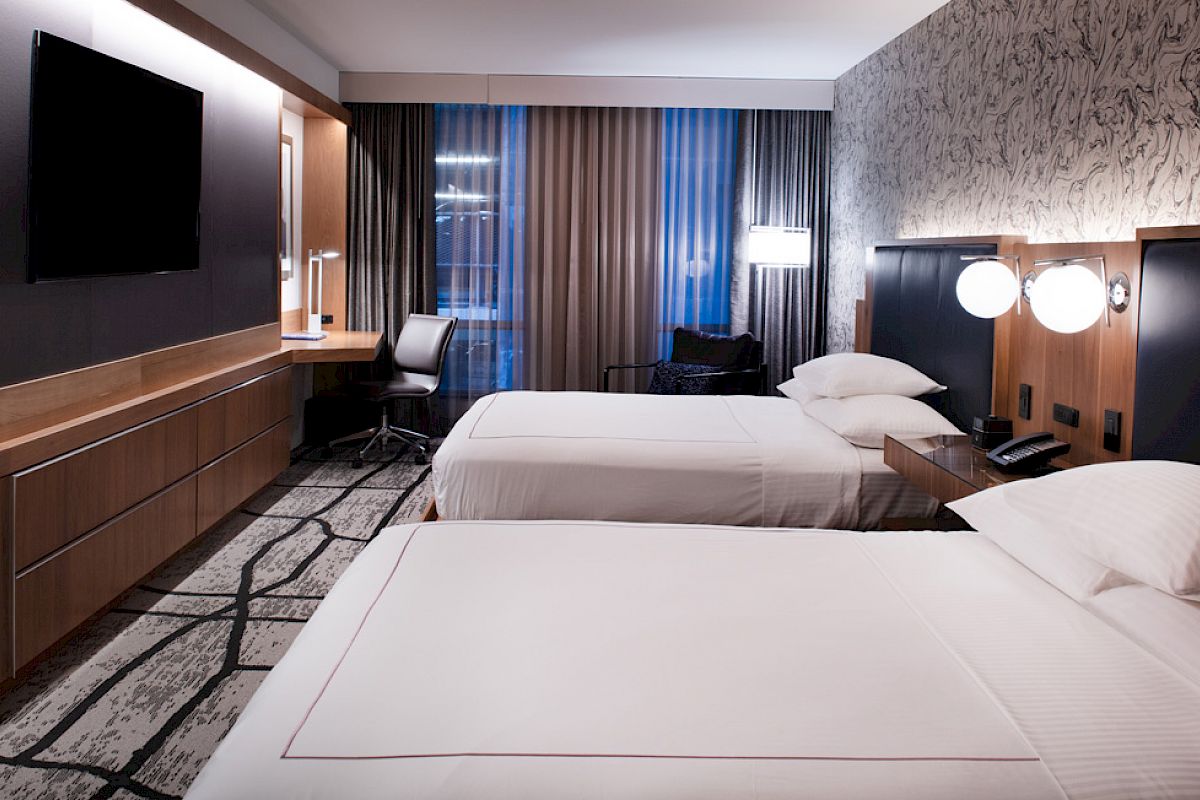 The image shows a modern hotel room with two beds, a TV, a desk, and ambient lighting, creating a cozy atmosphere.