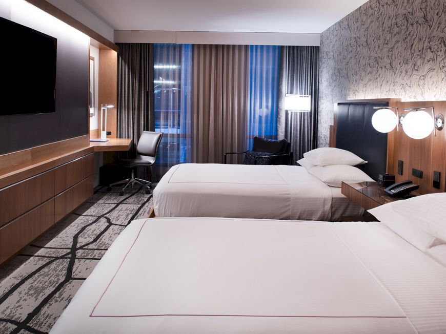 The image shows a modern hotel room with two beds, a TV, a desk, and ambient lighting, creating a cozy atmosphere.