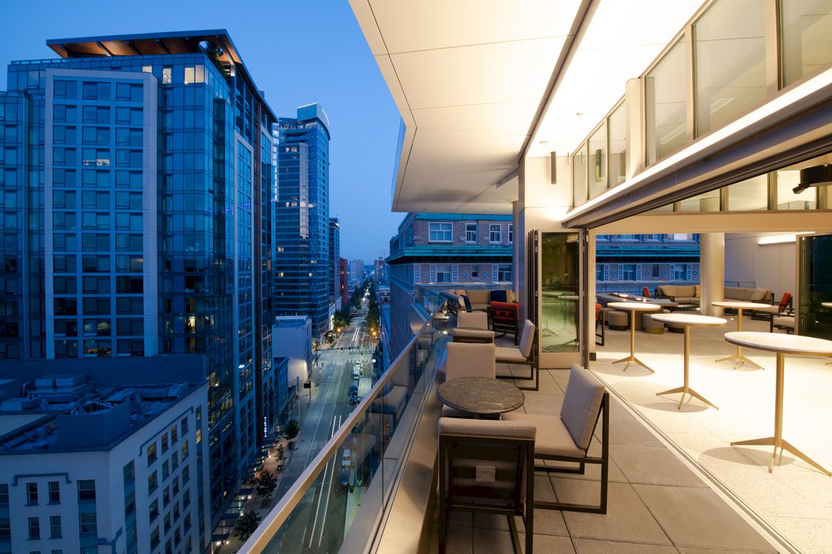 A modern balcony overlooking a cityscape at dusk, with tables and chairs set for dining and tall buildings visible in the background.