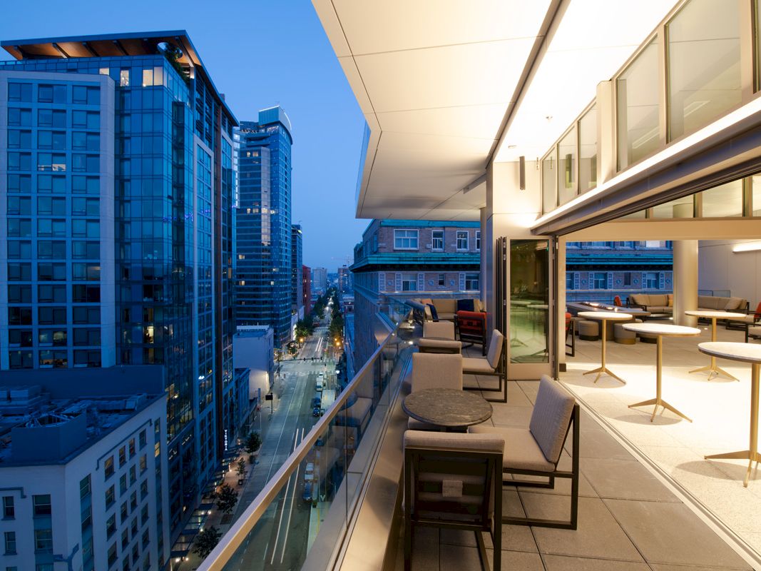 A modern balcony overlooking a cityscape at dusk, with tables and chairs set for dining and tall buildings visible in the background.