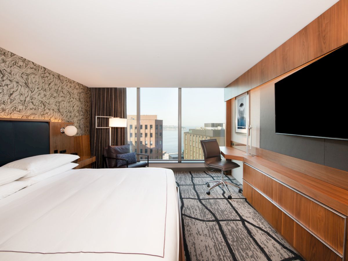 A modern hotel room with a large bed, wall-mounted TV, desk, chair, and a window view of buildings outside.