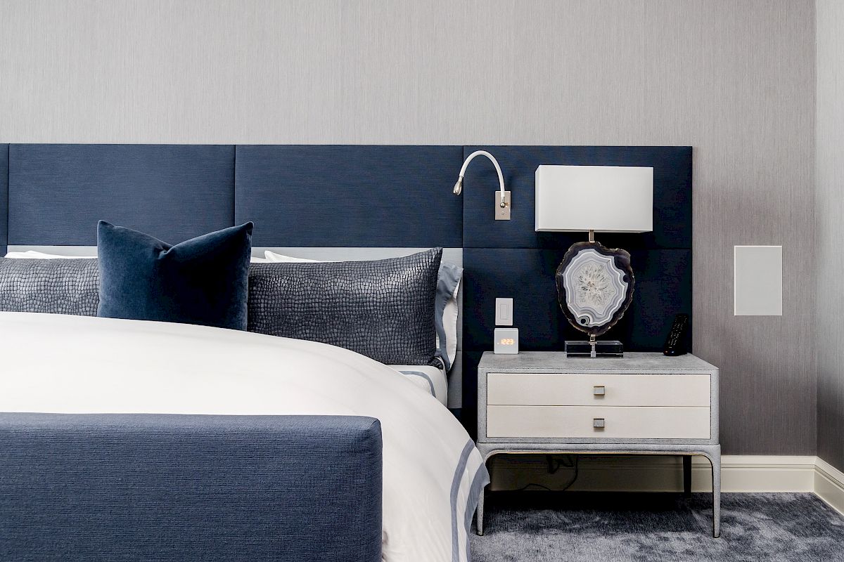 A modern bedroom with a bed, blue headboard, white nightstand, lamp, and decorative items on a plush carpet.