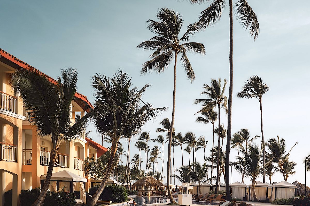 The image shows a tropical resort with palm trees, a pool, lounge chairs, and a building with balconies under a clear sky.
