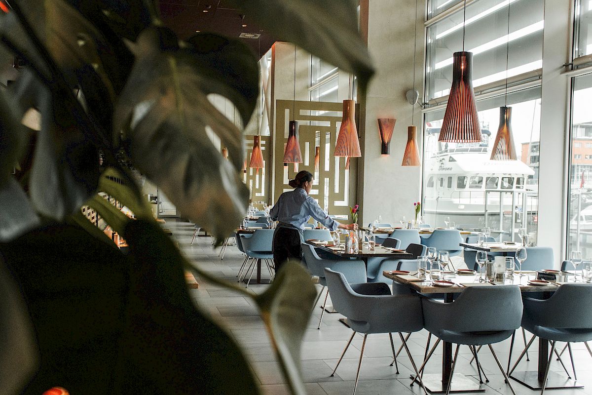 A person setting tables in a stylish, modern restaurant with large windows and unique pendant lights.