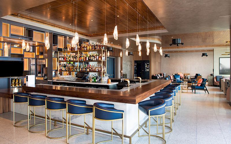 The image shows a modern bar with a central counter, stylish bar stools, and hanging lights in a spacious room.