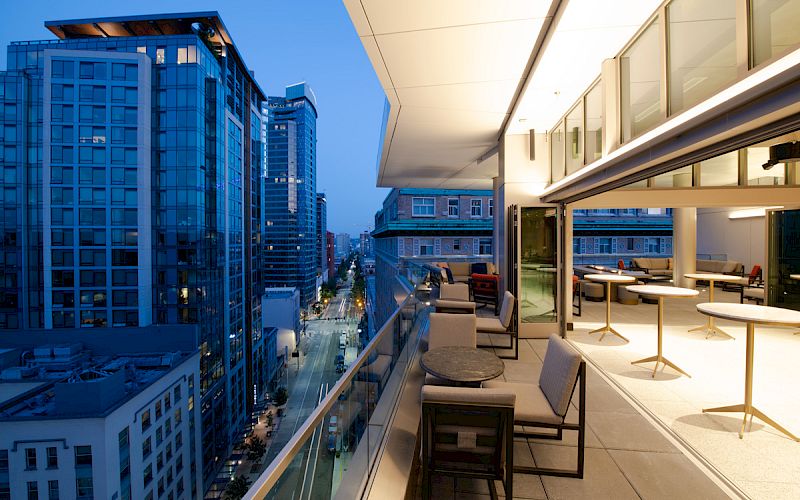 A modern balcony overlooking a cityscape with high-rise buildings is shown at dusk, featuring outdoor seating and tables under ambient lighting.